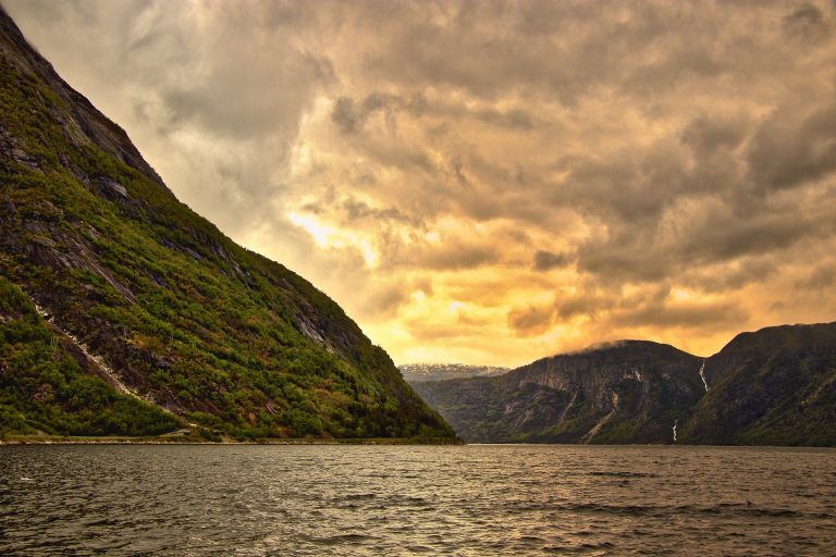 Ein Blick vom Wasser aus in die Richtung von Bergen, welche zum Teil grün bewachsen sind. Es ist bewölkt und dahinter scheint die Sonne.